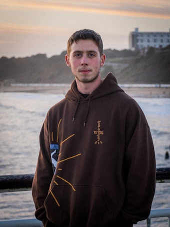 A person stands in front of a scenic background featuring the ocean and a coastal landscape. They are wearing a dark hoodie with a unique design on the front. The sky has a warm, pastel color with a building visible in the distance.