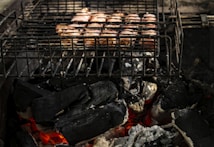 A grill laden with pieces of meat is positioned over glowing charcoal embers. The metal grid of the grill is darkened and contrasts with the vibrant orange and red hues of the burning coals beneath.
