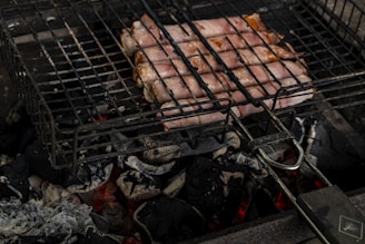 Slices of bacon wrapped around sausages are grilling on a barbecue over glowing charcoal. The cooking process is taking place on a metal grill rack positioned above hot coals and ash. The scene captures the raw and rustic essence of outdoor grilling.