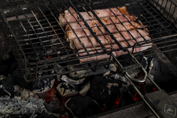 Slices of bacon wrapped around sausages are grilling on a barbecue over glowing charcoal. The cooking process is taking place on a metal grill rack positioned above hot coals and ash. The scene captures the raw and rustic essence of outdoor grilling.