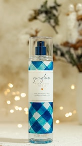 A bottle of fine fragrance mist with a blue checkered pattern on the label stands against a softly blurred background featuring bokeh lights and faint, indistinct foliage. The label reads 'gingham' by Bath & Body Works.