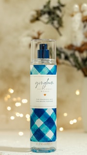 A bottle of fine fragrance mist with a blue checkered pattern on the label stands against a softly blurred background featuring bokeh lights and faint, indistinct foliage. The label reads 'gingham' by Bath & Body Works.