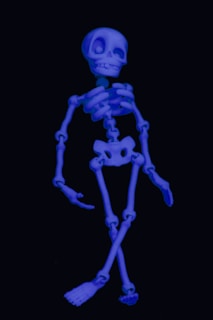 A cartoonish skeleton depicted in a glowing blue hue against a dark background. It has exaggerated, comical features with a large skull and oversized eye sockets, giving it a playful appearance.