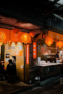 A cozy night market stall with orange lanterns hanging above, creating a warm glow. Two people are seated at a table outside, one of them is using a smartphone. A vendor stands by a counter preparing food. The signage and decor suggest a traditional and inviting street food atmosphere.