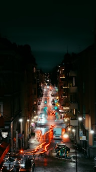 A nighttime urban street scene captures the vibrant energy of the city with glowing lights and streaking car trails. Multistory buildings line the street, their windows illuminated, adding a warm glow to the otherwise dark night. Wet pavement suggests recent rain, reflecting the lights and colors beautifully.