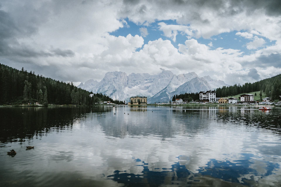 The image depicts a serene lake nestled amidst a mountainous landscape. The calm, reflective waters mirror the surrounding scenery, creating a beautiful symmetry. On one side of the lake, there is a small island with a white building on it, possibly a house or a cabin. In the distance, several houses can be seen scattered across the mountainside, adding to the picturesque setting. The sky above is filled with clouds, casting a soft light over the entire scene and enhancing its natural beauty.