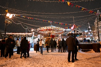 A lively winter fair with people gathered around a carousel surrounded by festive lights and decorations. Snow is falling gently, adding to the holiday atmosphere. Colorful flags and lights are strung overhead, and a Christmas tree is visible in the background.