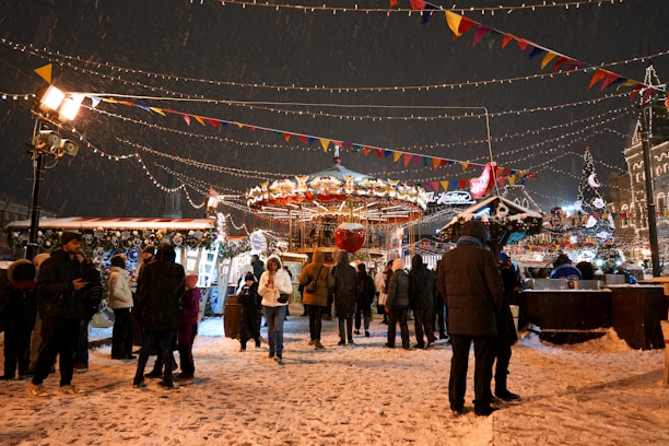A lively winter fair with people gathered around a carousel surrounded by festive lights and decorations. Snow is falling gently, adding to the holiday atmosphere. Colorful flags and lights are strung overhead, and a Christmas tree is visible in the background.