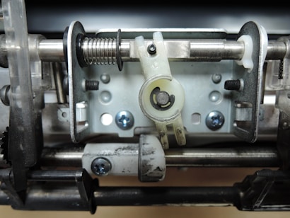 A close-up view of a mechanical component featuring metal rods, springs, and brackets. The assembly includes screws and a plastic piece with a circular cutout positioned centrally. The components are arranged in a structural and functional layout, indicative of machinery or an electronic device.