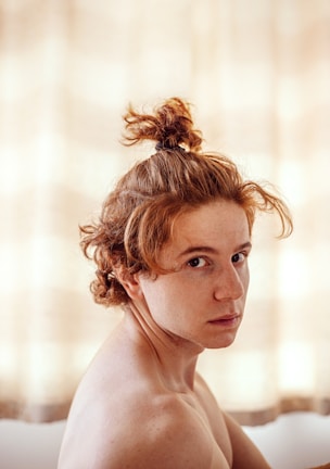 A person with light skin tone and red hair styled in a small top knot looks towards the camera. The background is softly lit, creating a warm and intimate atmosphere.