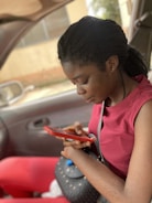 A person sitting in a car, engrossed in their mobile phone. They are wearing a red top and holding a red phone, with a black purse resting on their lap.