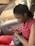 A person sitting in a car, engrossed in their mobile phone. They are wearing a red top and holding a red phone, with a black purse resting on their lap.