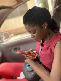 A person sitting in a car, engrossed in their mobile phone. They are wearing a red top and holding a red phone, with a black purse resting on their lap.