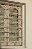 A window with ornate metal bars featuring symmetrical, decorative scroll patterns. The window appears to be closed with horizontal blinds partially visible behind the bars. The wall surrounding the window is painted in a light color and has a textured surface.