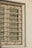 A window with ornate metal bars featuring symmetrical, decorative scroll patterns. The window appears to be closed with horizontal blinds partially visible behind the bars. The wall surrounding the window is painted in a light color and has a textured surface.