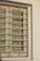 A window with ornate metal bars featuring symmetrical, decorative scroll patterns. The window appears to be closed with horizontal blinds partially visible behind the bars. The wall surrounding the window is painted in a light color and has a textured surface.