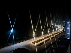 A night scene with an illuminated pathway lined with streetlights, casting star-like flares. The path is flanked by small bushes and a clear sky above enhances the glowing effect of the lights. The scene conveys a sense of tranquility and quietness.