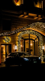 A luxurious car is parked in front of a grand entrance elaborately adorned with twinkling golden fairy lights. The architecture is classic with ornate details, and warm light emanates from the windows and lanterns by the door. Green wreaths add a touch of festive decor to the scene.