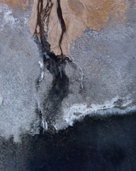 An aerial view of a landscape with intricate patterns and textures. Light earthy tones blend into darker areas, indicating possible riverbeds or erosion lines on a rugged terrain.