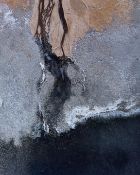 An aerial view of a landscape with intricate patterns and textures. Light earthy tones blend into darker areas, indicating possible riverbeds or erosion lines on a rugged terrain.