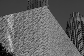 A modern architectural structure with a facade covered in diagonal marble tiles. The building is juxtaposed with classic skyscrapers in the background, showcasing a mix of contemporary and traditional design elements. The image is in black and white, highlighting the texture and patterns of the marble.