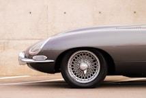 A vintage car's front section showcasing sleek curves and a bright metallic finish. The focus is on the intricate design of the wheel, which features a classic wire-spoke pattern. The chrome details add a touch of elegance against a neutral concrete background.
