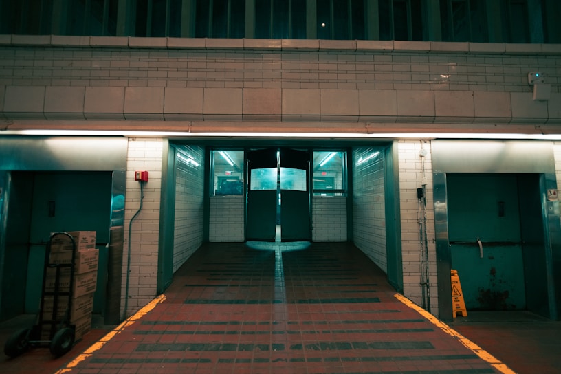 A hallway with industrial design featuring tiled walls and dim lighting. There are two large elevator doors on either side, a stack of boxes on a dolly to the left, and a wet floor caution sign on the right. The floor is made of red tiles, and fluorescent lighting casts a greenish hue throughout the area.