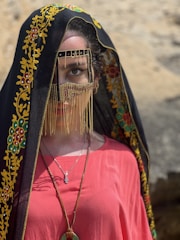 A person wearing a traditional garment with intricate embroidery and vibrant colors. The headpiece includes a black veil with yellow and red floral patterns, covering the head and parts of the face. A decorative mask with golden chains partially conceals the face, highlighting one eye. The person wears a vivid red top and a necklace with a pendant.