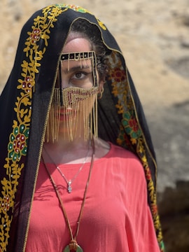A person wearing a traditional garment with intricate embroidery and vibrant colors. The headpiece includes a black veil with yellow and red floral patterns, covering the head and parts of the face. A decorative mask with golden chains partially conceals the face, highlighting one eye. The person wears a vivid red top and a necklace with a pendant.