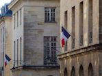 A street view of a building facade with a beige stone exterior. French flags are displayed on metal poles extending from the building at two locations. The windows are rectangular with grid patterns. Decorative street lamps are also attached to the building.