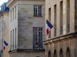 A street view of a building facade with a beige stone exterior. French flags are displayed on metal poles extending from the building at two locations. The windows are rectangular with grid patterns. Decorative street lamps are also attached to the building.