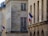 A street view of a building facade with a beige stone exterior. French flags are displayed on metal poles extending from the building at two locations. The windows are rectangular with grid patterns. Decorative street lamps are also attached to the building.