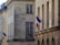 A street view of a building facade with a beige stone exterior. French flags are displayed on metal poles extending from the building at two locations. The windows are rectangular with grid patterns. Decorative street lamps are also attached to the building.