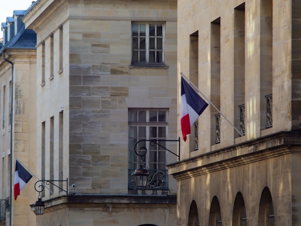 A street view of a building facade with a beige stone exterior. French flags are displayed on metal poles extending from the building at two locations. The windows are rectangular with grid patterns. Decorative street lamps are also attached to the building.