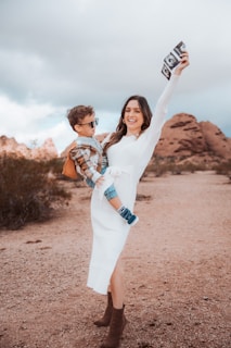 A woman in a white dress stands on a desert path, holding ultrasound images and a young boy wearing sunglasses. They appear cheerful, with the boy dressed in a plaid shirt and denim shorts. The landscape features rocky formations and cloudy skies.