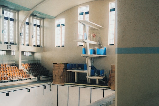 An indoor swimming pool with a large diving platform against a wall with tall, narrow windows. The walls and ceilings feature a smooth, light-colored finish that contrasts with the darker seating area made up of wooden bleachers. The diving platform includes multiple levels and a ladder, and the pool itself is empty.