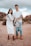 A family standing outdoors in a desert-like setting with a rocky backdrop. The woman is wearing a white dress and holding ultrasound images, while the man in a white shirt and ripped jeans is holding a child by the arms, playfully suspending them in front.