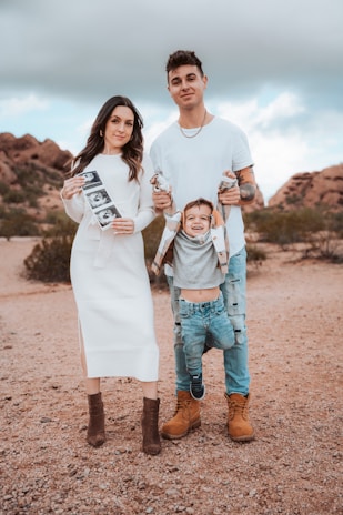 A family standing outdoors in a desert-like setting with a rocky backdrop. The woman is wearing a white dress and holding ultrasound images, while the man in a white shirt and ripped jeans is holding a child by the arms, playfully suspending them in front.