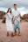 A family standing outdoors in a desert-like setting with a rocky backdrop. The woman is wearing a white dress and holding ultrasound images, while the man in a white shirt and ripped jeans is holding a child by the arms, playfully suspending them in front.