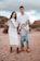 A family standing outdoors in a desert-like setting with a rocky backdrop. The woman is wearing a white dress and holding ultrasound images, while the man in a white shirt and ripped jeans is holding a child by the arms, playfully suspending them in front.