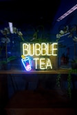 A neon sign reading 'Bubble Tea' with an image of a cup with a straw and bubbles is illuminated in a dark setting. The background includes reflections, potted plants, and a partially visible room with some furniture and additional decor.