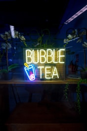 A neon sign reading 'Bubble Tea' with an image of a cup with a straw and bubbles is illuminated in a dark setting. The background includes reflections, potted plants, and a partially visible room with some furniture and additional decor.