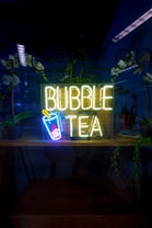 A neon sign reading 'Bubble Tea' with an image of a cup with a straw and bubbles is illuminated in a dark setting. The background includes reflections, potted plants, and a partially visible room with some furniture and additional decor.