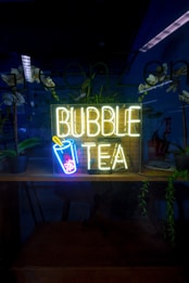 A neon sign reading 'Bubble Tea' with an image of a cup with a straw and bubbles is illuminated in a dark setting. The background includes reflections, potted plants, and a partially visible room with some furniture and additional decor.