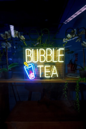 A neon sign reading 'Bubble Tea' with an image of a cup with a straw and bubbles is illuminated in a dark setting. The background includes reflections, potted plants, and a partially visible room with some furniture and additional decor.