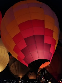 A large, colorful hot air balloon with a pattern of red, pink, orange, and yellow segments is illuminated against a dark background. Other balloons are partially visible in the background, suggesting a nighttime balloon festival.