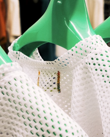 A green clothes hanger holds a perforated white garment with textured material. Slight colorful stitching is visible on the fabric, which features numerous small holes.
