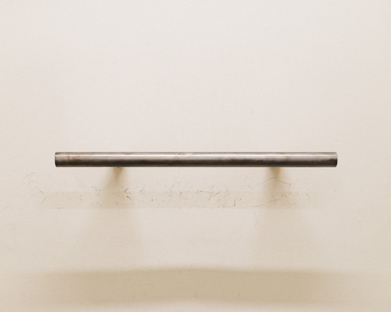 A minimalist scene featuring a single metallic horizontal bar mounted on a plain, off-white wall. The bar casts a soft shadow that blends into the subtly textured surface beneath. There are faint, irregular marks around the mounting area on the wall, adding a hint of imperfection to the composition.