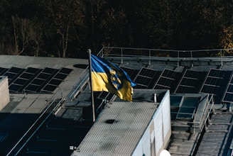 A flag with yellow and blue colors featuring a coat of arms is prominently displayed on a rooftop equipped with solar panels. The rooftop is surrounded by trees, indicating a mix of natural and industrial elements.
