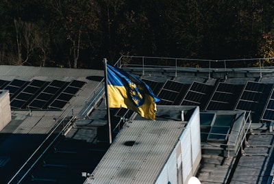 A flag with yellow and blue colors featuring a coat of arms is prominently displayed on a rooftop equipped with solar panels. The rooftop is surrounded by trees, indicating a mix of natural and industrial elements.